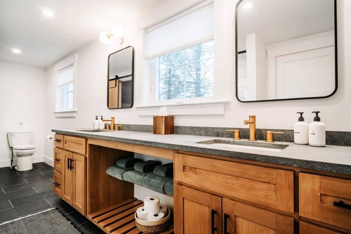 Modern farmhouse bathroom featuring a double vanity with light wood cabinets, dark stone countertops, gold fixtures, and two rectangular mirrors, illuminated by natural light from a large window.