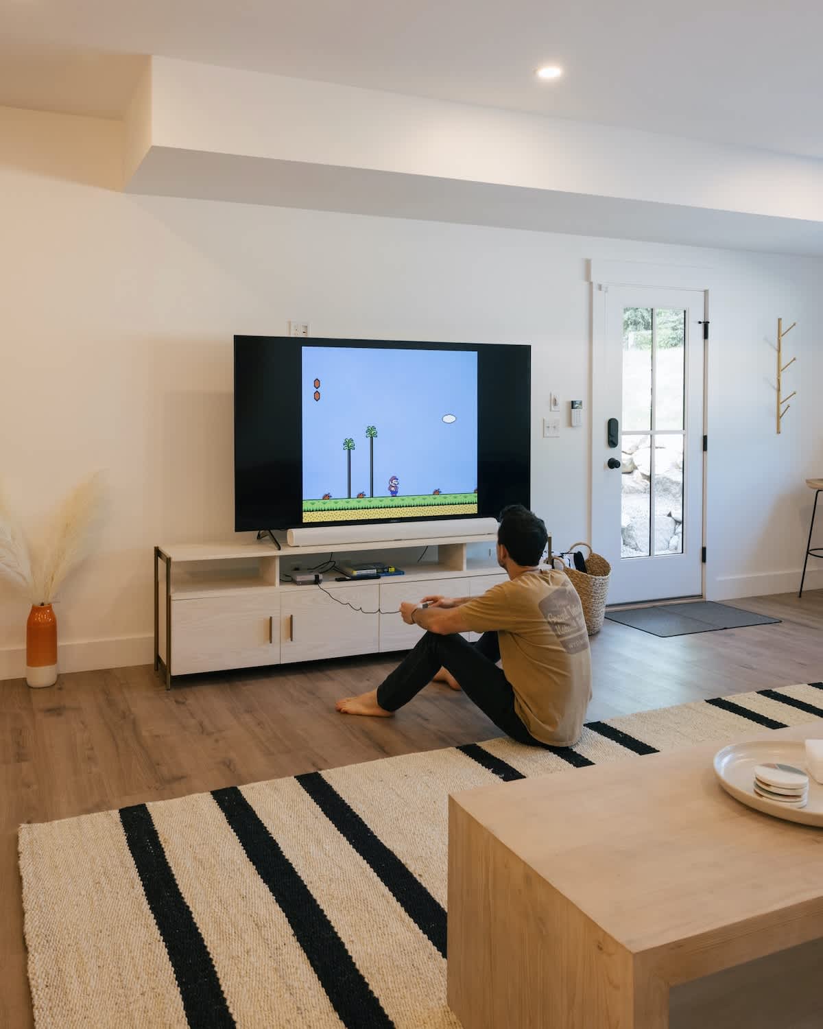 Cozy recreation space with a large TV displaying a video game, where a person sits on the floor playing. The room features light wood flooring, a striped area rug, and a white media console.