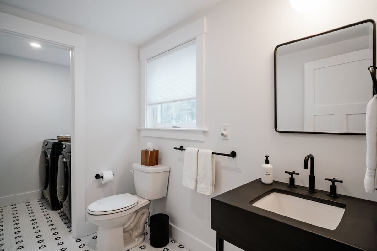 Modern bathroom with a black vanity, white sink, and black fixtures, featuring a large rectangular mirror. A toilet is next to it, and a laundry area with a washer and dryer is visible through an open doorway.