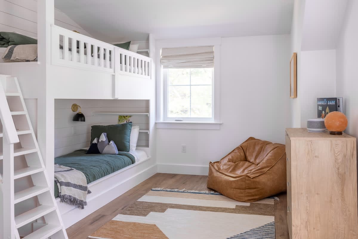 Children's bedroom featuring custom white built-in bunk beds with ladders, a cozy brown bean bag chair, a patterned rug, and a window with a Roman shade, all on light wood flooring.
