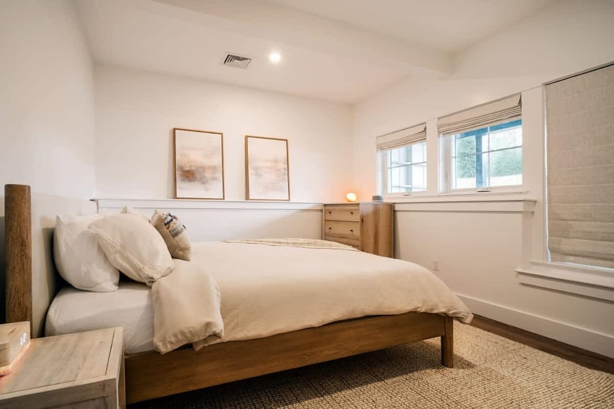A cozy bedroom features a light wooden bed frame with neutral bedding and pillows. Two abstract art pieces hang above the headboard, and a wooden dresser sits beside a window with Roman shades, creating a serene atmosphere.
