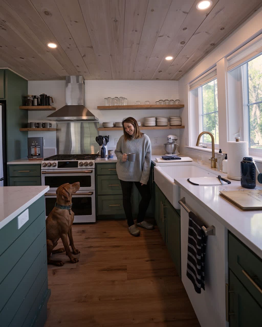 Bright kitchen with sage green shaker cabinets, white countertops, and open wood shelves. A woman stands by the farmhouse sink with a dog sitting on the wood floor, showcasing a gold faucet.