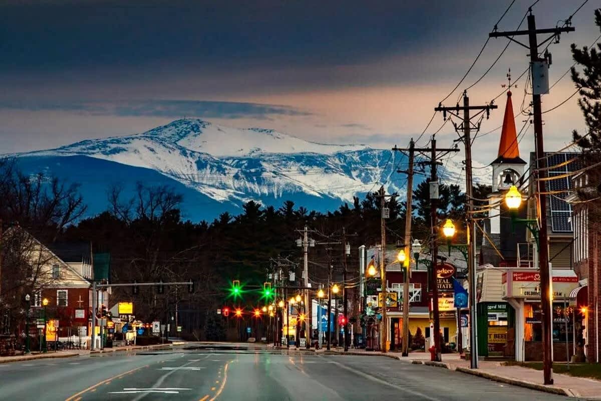 A picturesque town street at dusk, illuminated by warm streetlights, with power lines overhead and a majestic snow-capped mountain range in the distant background under a dramatic sky.