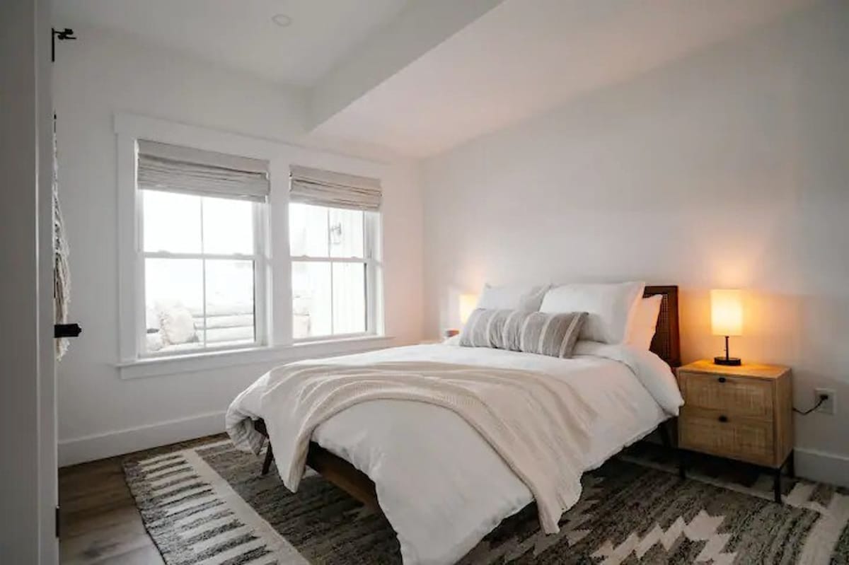 A bright bedroom offers a comfortable bed with white bedding and a striped throw blanket. A rustic wooden nightstand with a lamp is next to the bed, and a patterned rug covers the hardwood floor, illuminated by natural light from two windows.