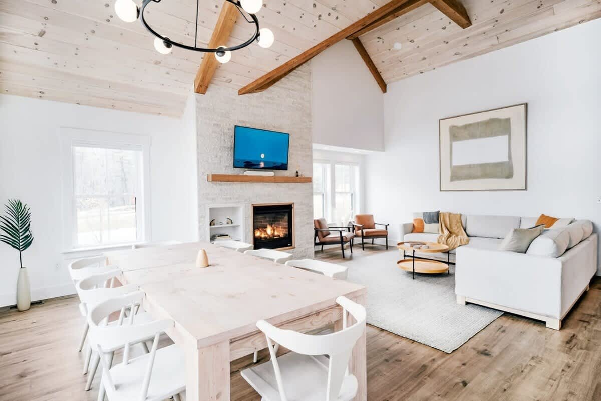 Spacious open-concept living and dining area with vaulted wood-paneled ceilings and exposed wood beams. A large light wood dining table with white chairs is in the foreground, leading to a living space with a white sofa and stone fireplace.