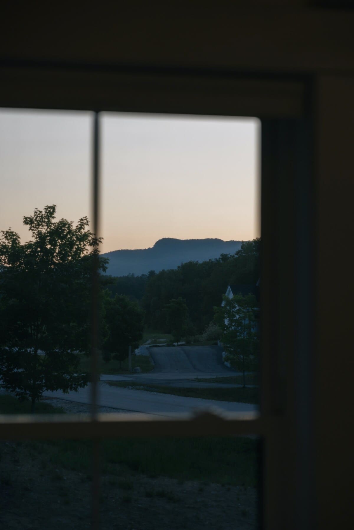 Serene view through a window at dusk, showing a winding road, lush green trees, and distant mountains under a soft, fading sky.