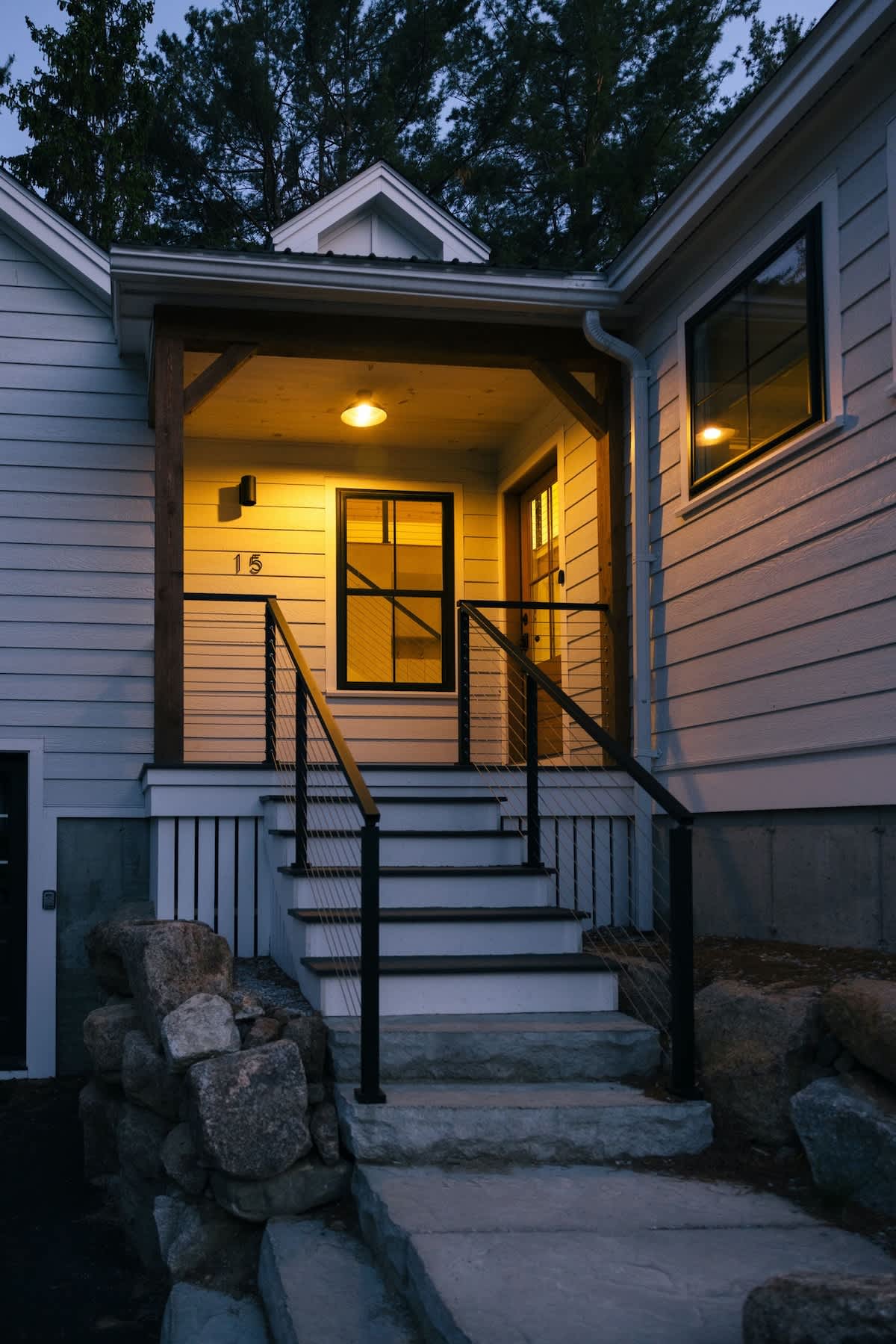 The illuminated front entrance of the house at dusk, featuring stone steps leading up to a covered porch with wooden beams and black railings. A house number '15' is visible next to the door, with warm light glowing from inside.