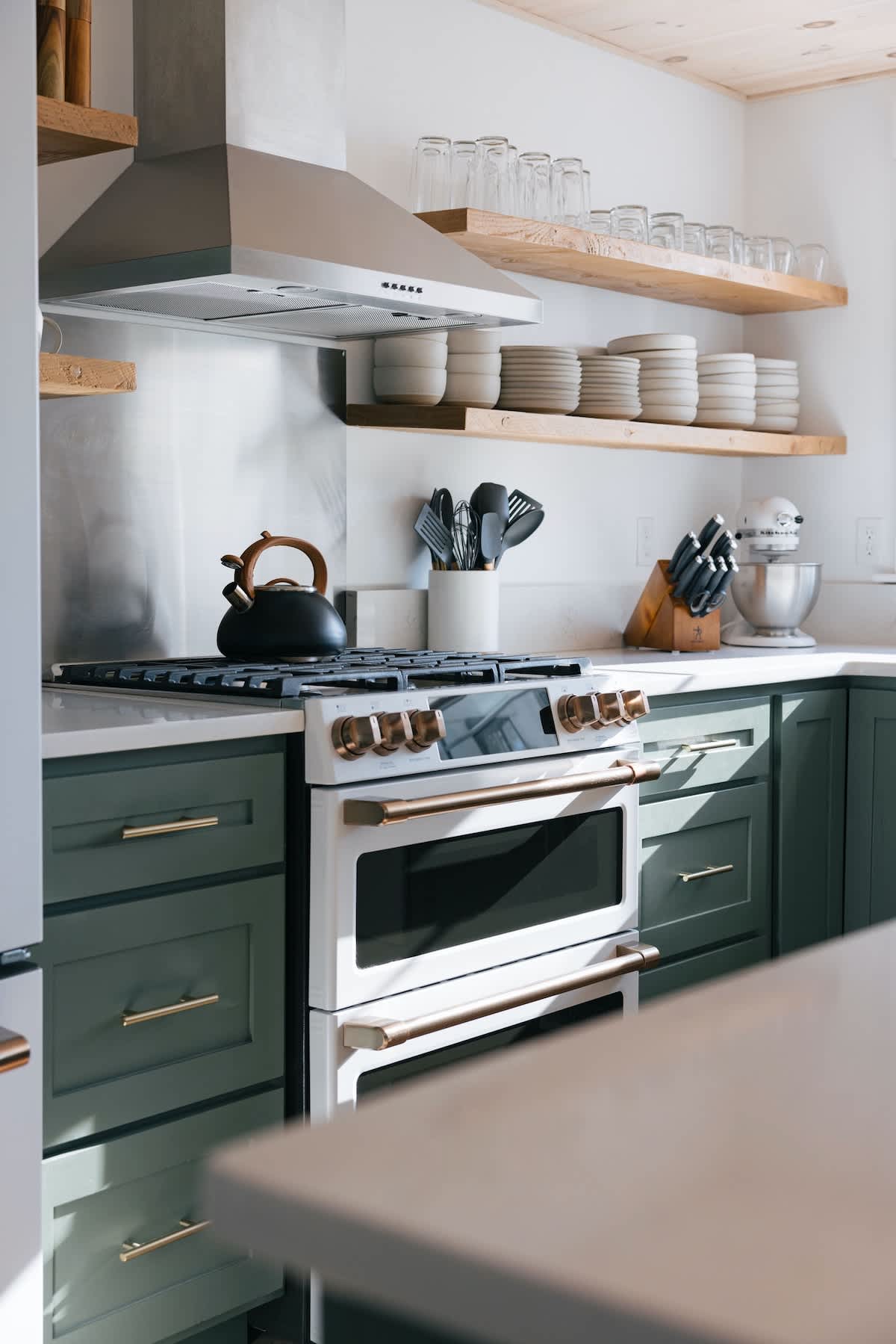 Modern farmhouse kitchen featuring sage green lower cabinets, white countertops, and open wooden shelving. A white double oven range with brass accents is central, topped by a stainless steel range hood.