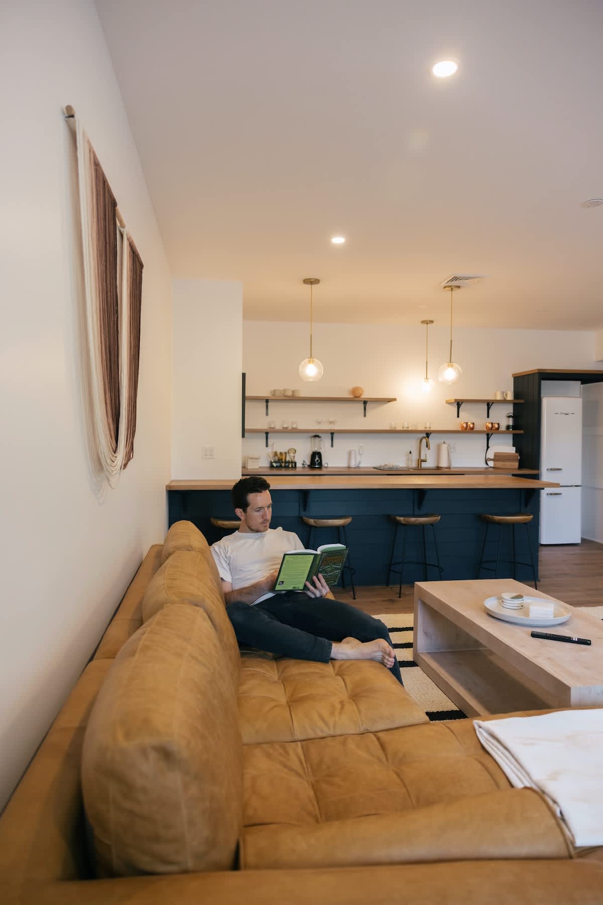 A man relaxes on a tufted brown leather sofa in an open-concept living area, reading a book. In the background, a modern wet bar with dark blue cabinets and open shelving is visible, connecting the spaces.