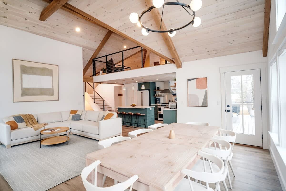 Expansive open-concept living and dining area featuring a vaulted wood-paneled ceiling with exposed beams, a comfortable sectional sofa, a large dining table, and a view into the kitchen and a loft area.