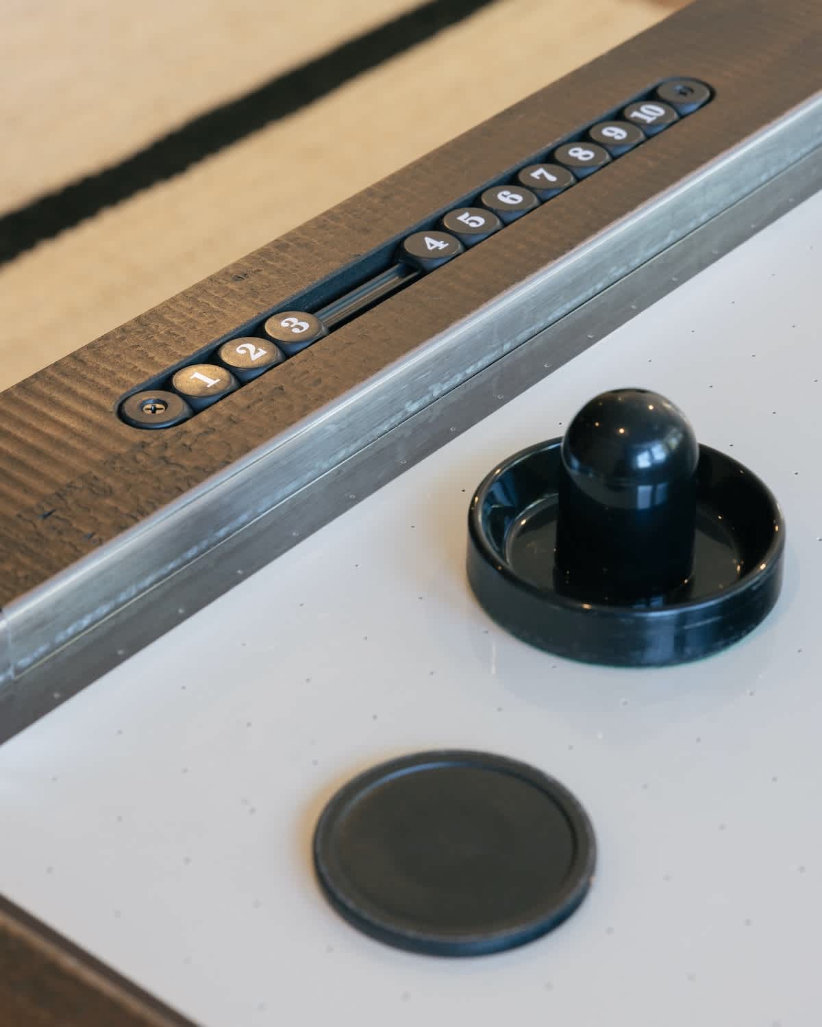 A close-up view highlights the scoring mechanism of an air hockey table, featuring numbered sliders and two black pucks on the white playing surface. The table has a rustic wooden frame.