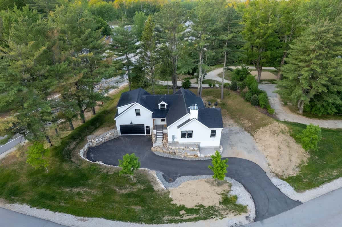 High aerial view of a modern white farmhouse with a black roof, nestled among lush green trees. A paved driveway curves around the front of the house, leading to a garage, with a small road visible in the distance.