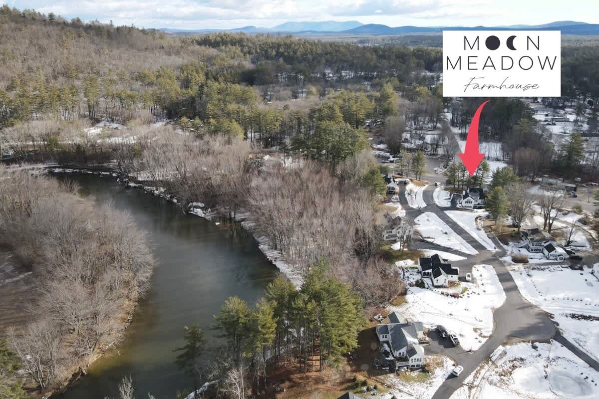 An aerial map view showing the 'Moon Meadow Farmhouse' logo and an arrow pointing to the property, situated near a winding river and surrounded by a snowy, wooded landscape. Other houses and roads are visible in the neighborhood.