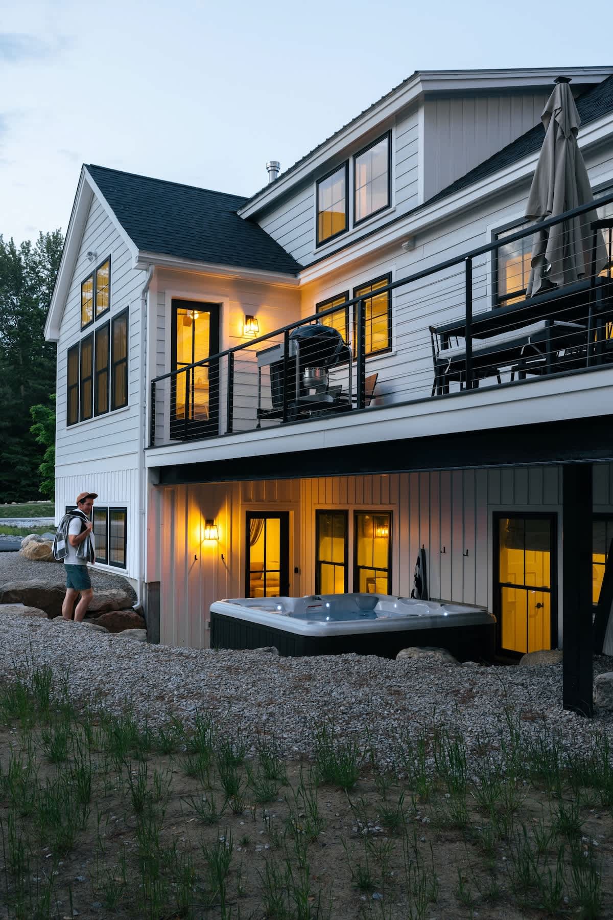 Modern white farmhouse exterior with multiple windows and a second-story deck, illuminated at dusk. A hot tub is visible on the ground level, surrounded by gravel and natural landscaping.