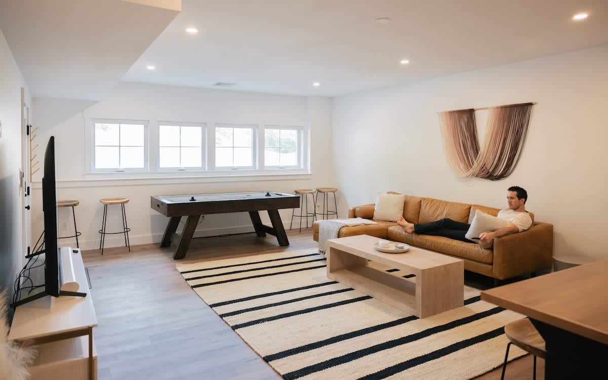 A bright game room features a large brown leather sofa, a light wood coffee table, and a black and white striped rug. An air hockey table sits near a row of windows, while a TV is mounted on the opposite wall, creating a versatile entertainment space.