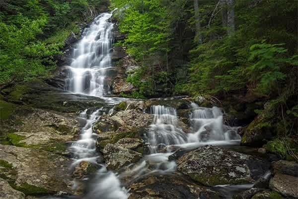 A beautiful multi-tiered waterfall cascades over mossy rocks into a clear stream, surrounded by vibrant green forest foliage, creating a serene natural landscape.