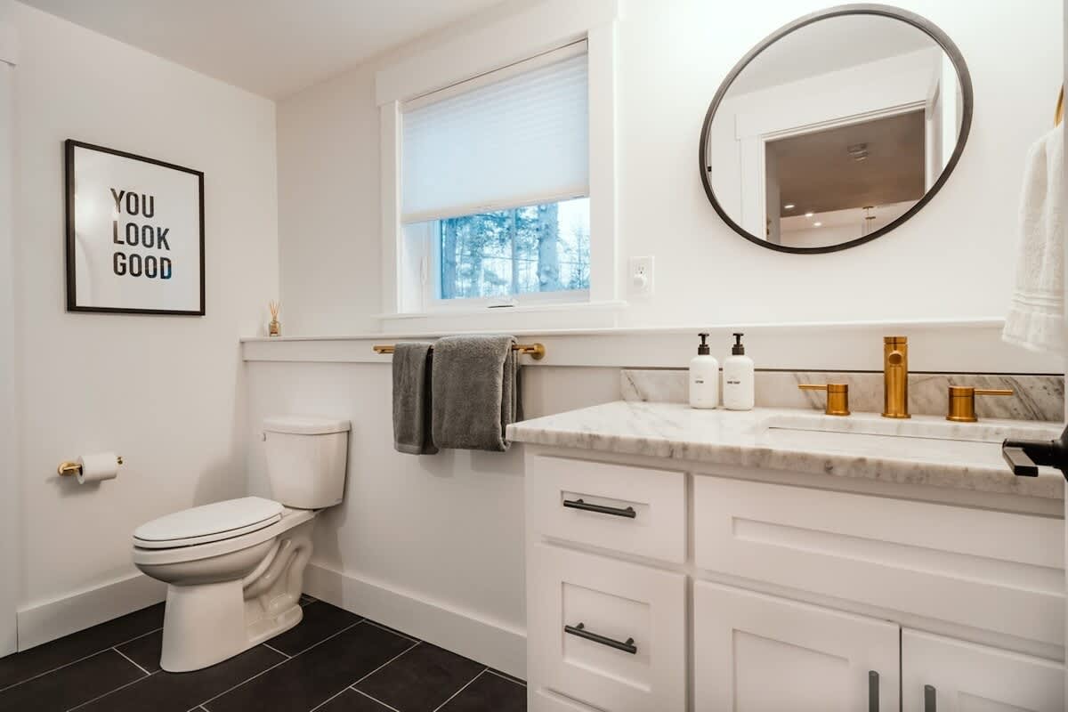 Modern white bathroom with dark tiled floor, a white vanity with marble countertop and gold fixtures, and a toilet. A black-framed mirror and a 'YOU LOOK GOOD' sign add stylish accents.