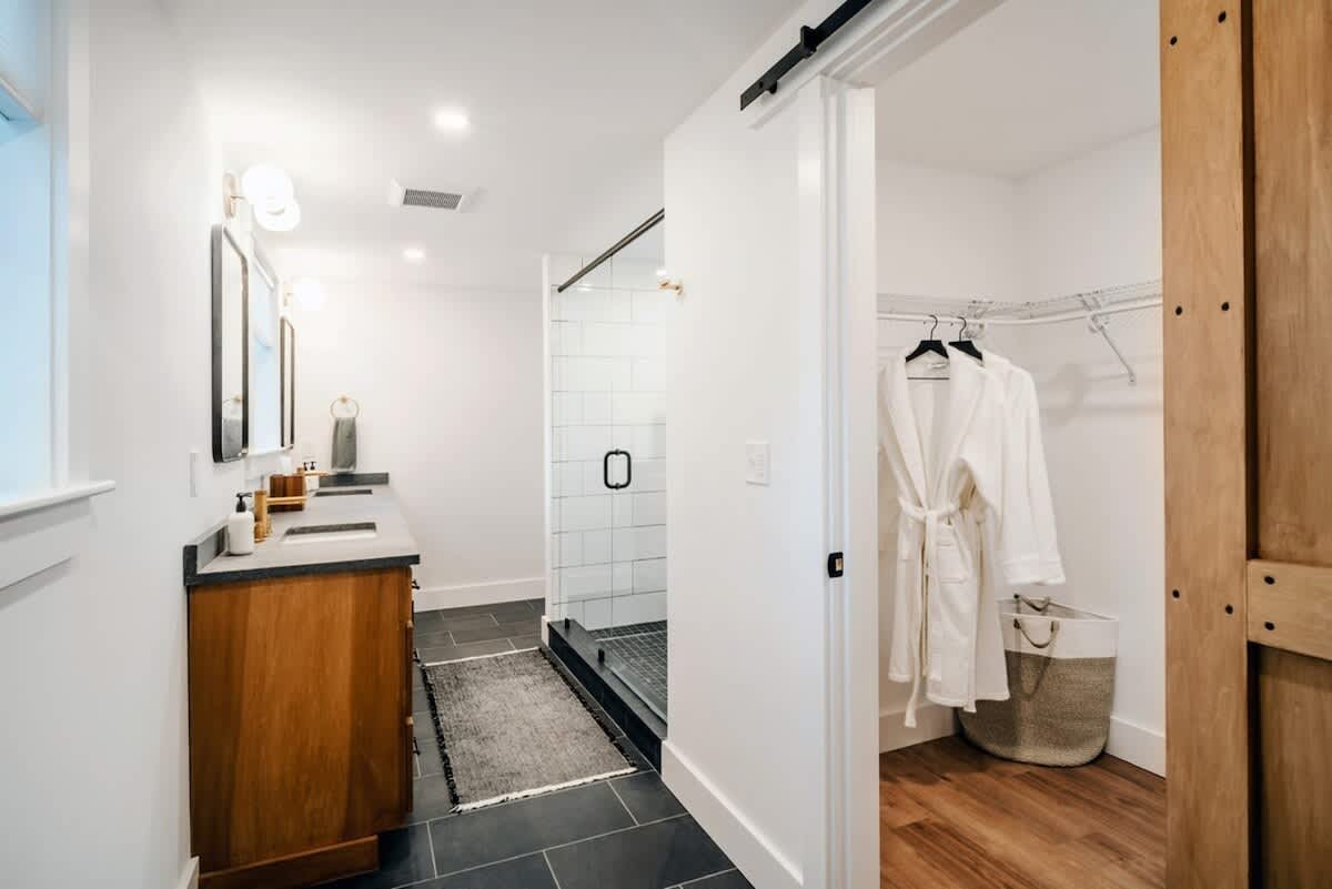 View of a modern farmhouse bathroom with a glass-enclosed shower featuring white subway tiles, a dark tiled floor, and an adjacent open closet with a white robe and laundry basket.