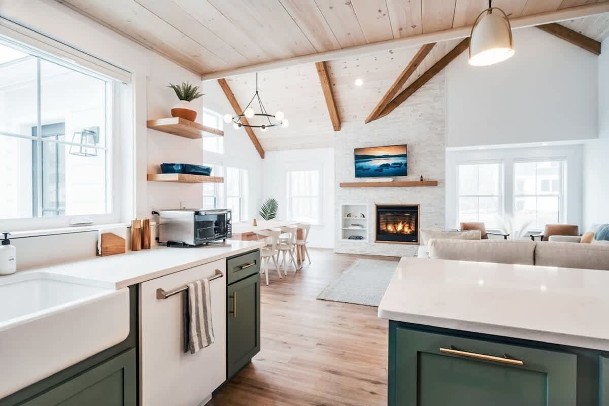 Open-concept living space with vaulted wooden ceilings and exposed beams, featuring a modern kitchen with dark green lower cabinets and a farmhouse sink. The dining area and living room with a fireplace are visible beyond.