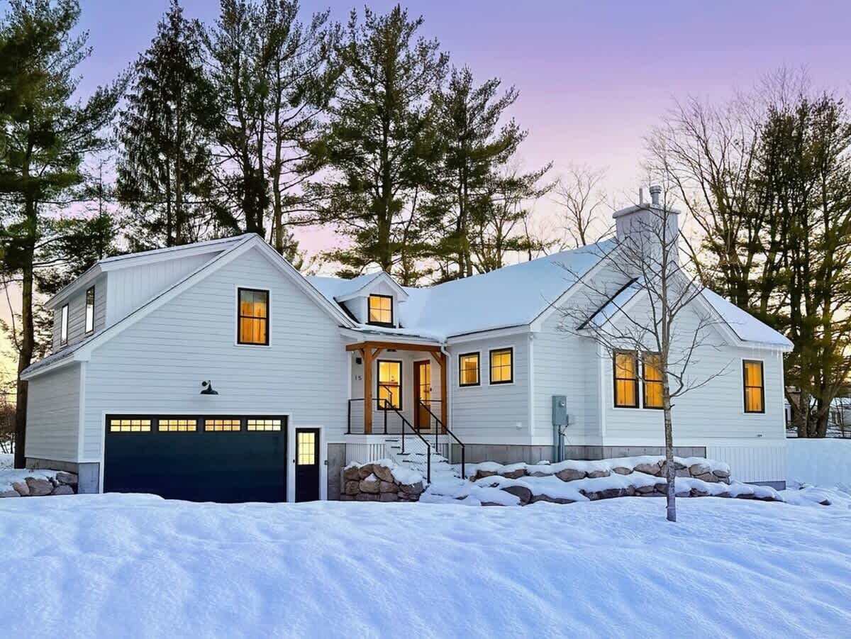 The front of a modern white farmhouse with a black roof, covered in snow under a soft twilight sky. It features a dark garage door, a covered entrance with wooden beams, and illuminated windows, surrounded by snow-covered landscaping.