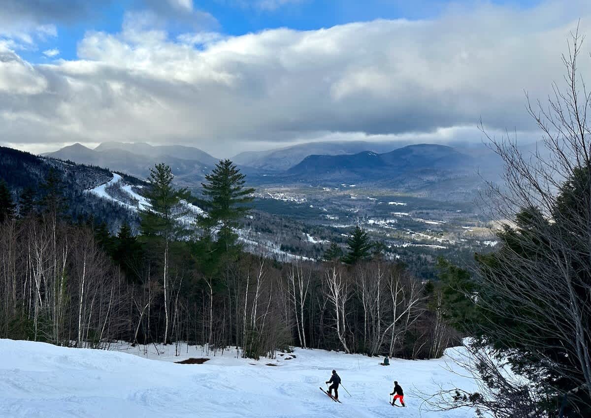 A panoramic view from a snowy ski slope, featuring two skiers descending with a vast valley and a distant town nestled among forested mountains under a dramatic cloudy sky.