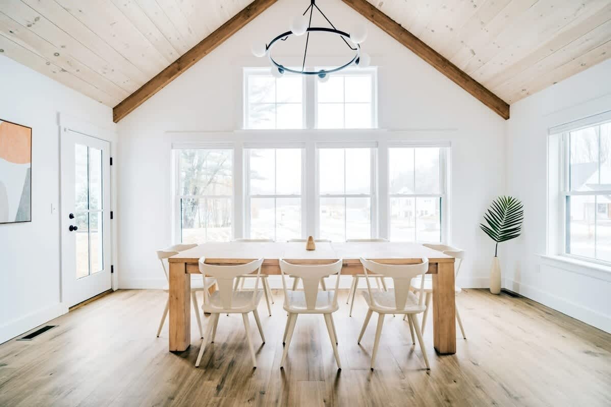 Spacious dining area with a long light wood table and white chairs, set under a vaulted wood-paneled ceiling with exposed beams, and surrounded by large windows offering ample natural light.
