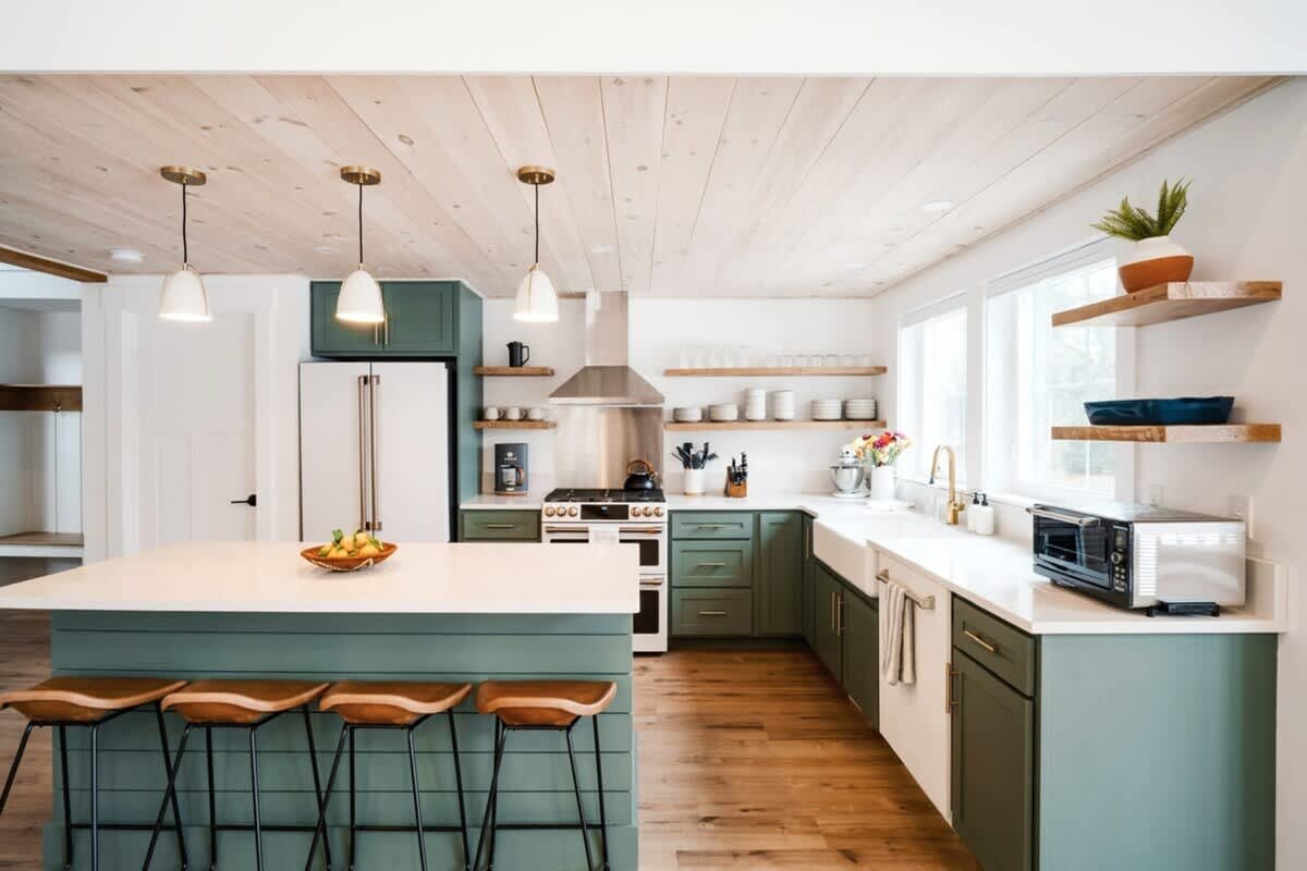 Wide view of a modern kitchen with sage green base cabinets, white countertops, a large island with bar seating, open shelving, and a light wood-paneled ceiling.