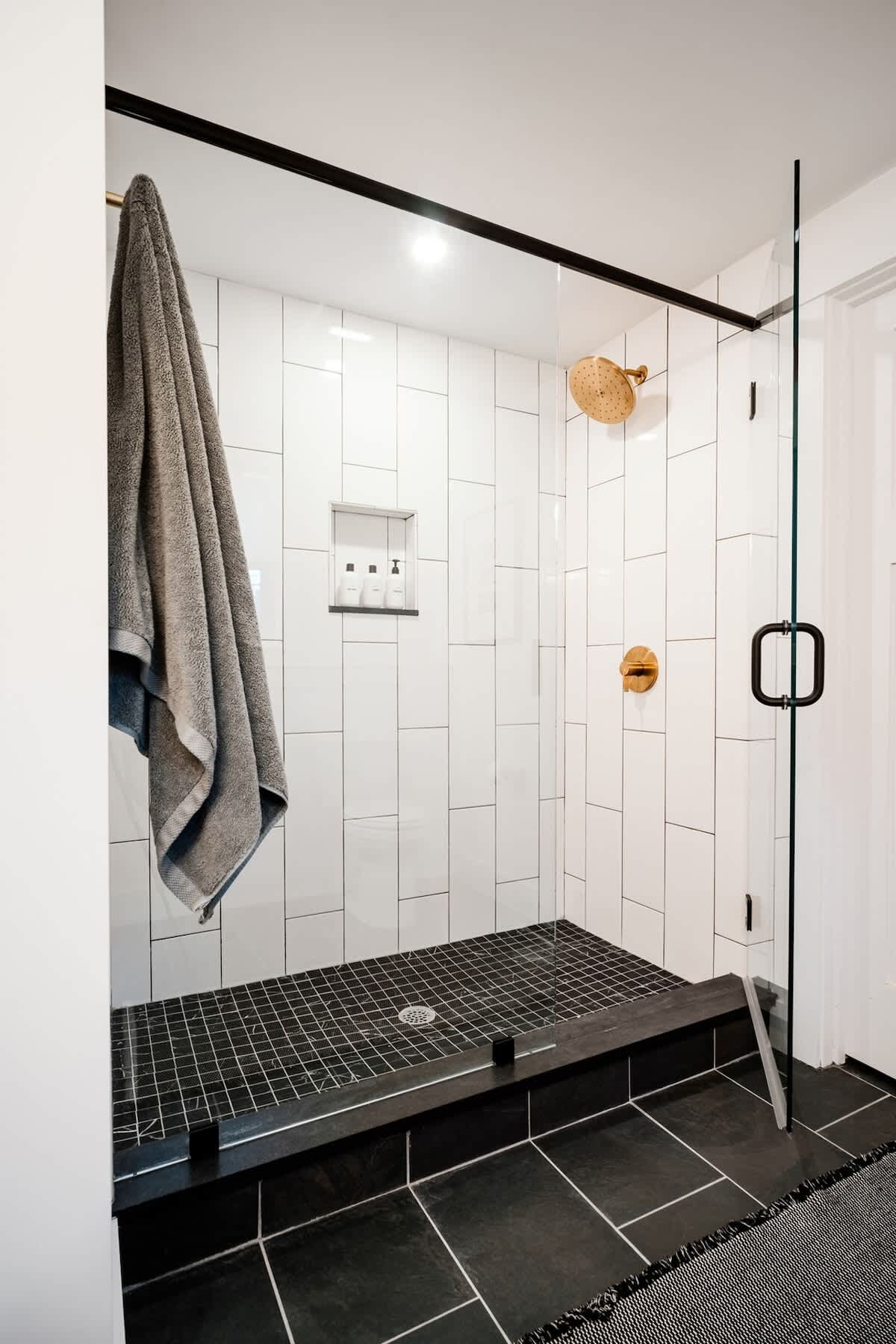 A modern walk-in shower features white subway tiles on the walls and small black mosaic tiles on the shower floor. A black-framed glass enclosure, a gold shower head, and a matching faucet complete the sleek design.