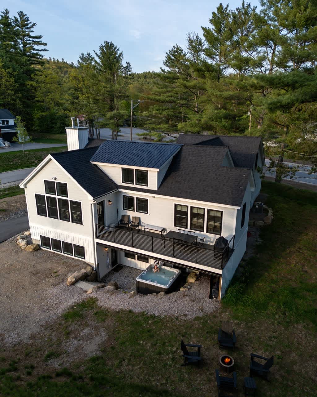 Aerial view of a modern white farmhouse with a black roof, featuring a large deck with outdoor seating and a hot tub on the lower level. A fire pit with Adirondack chairs is set on a gravel patch, surrounded by tall pine trees.