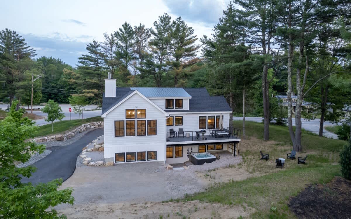 Aerial view of a contemporary white farmhouse with a black roof, showcasing a spacious deck with outdoor furniture and a hot tub below. The property is surrounded by tall pine trees, with a paved driveway and road visible in the distance.