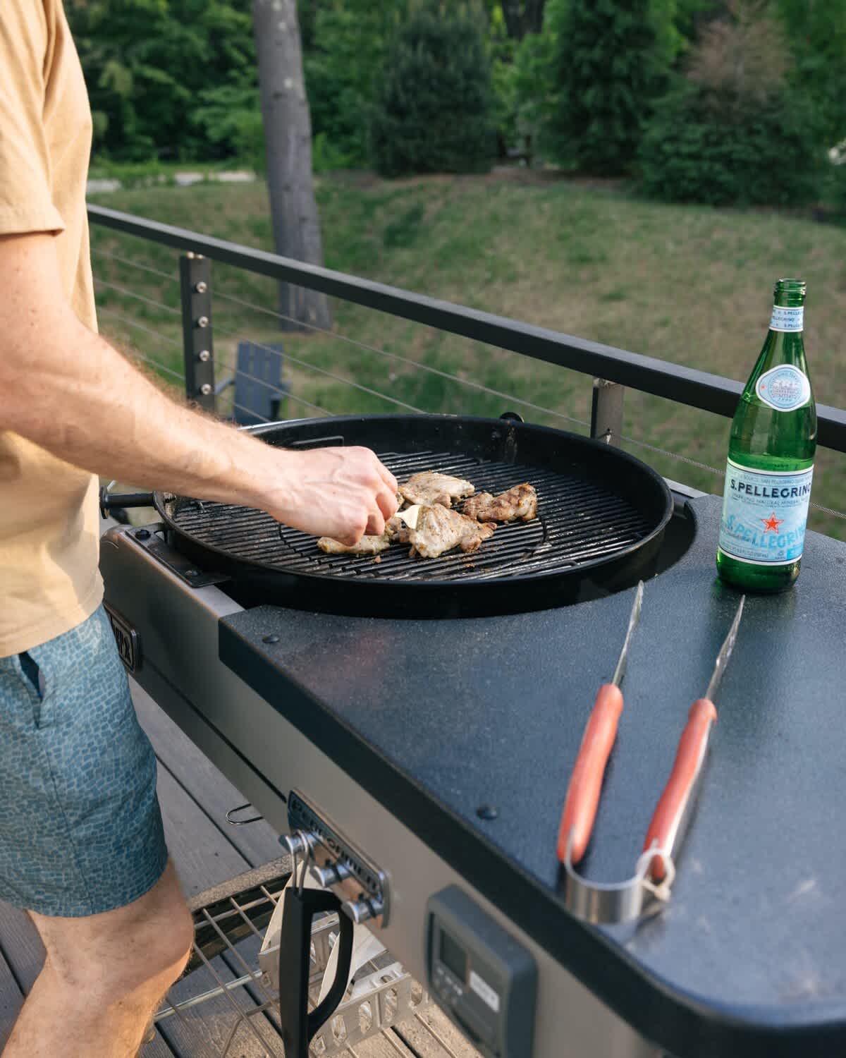 Close-up of a person grilling meat on a modern outdoor grill on a deck. A bottle of sparkling water and grilling tongs are on the counter next to the grill.