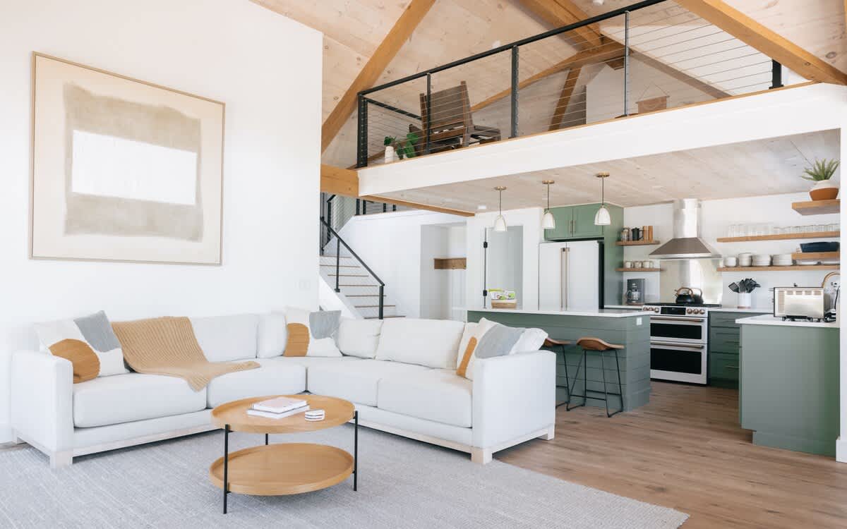 Spacious open-concept living area with a white sectional sofa, two round wooden coffee tables, a light rug, and an adjacent kitchen featuring sage green cabinets, a large island, and vaulted ceilings with exposed beams.