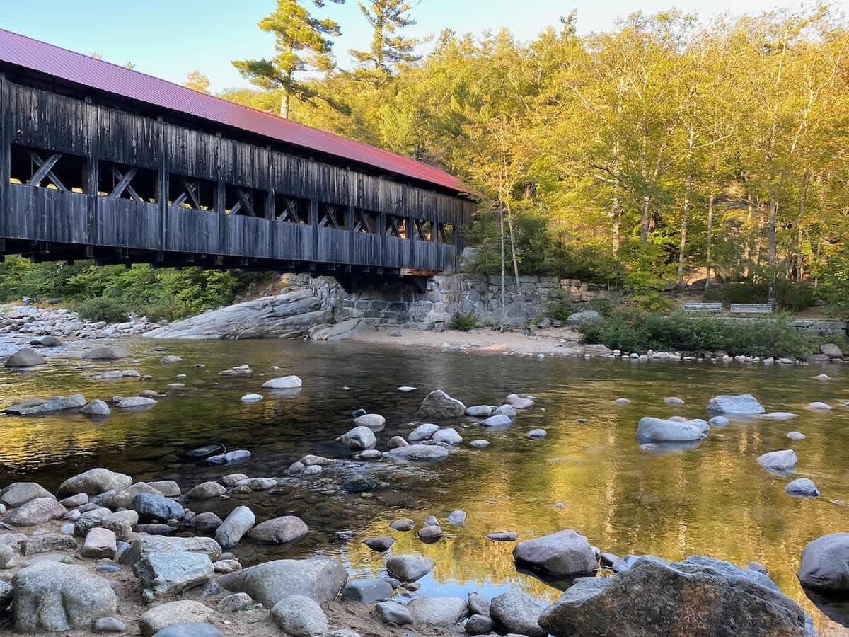 A classic wooden covered bridge with a red roof spans a clear, rocky river, surrounded by lush green and golden autumn foliage under a bright sky, reflecting in the calm water.
