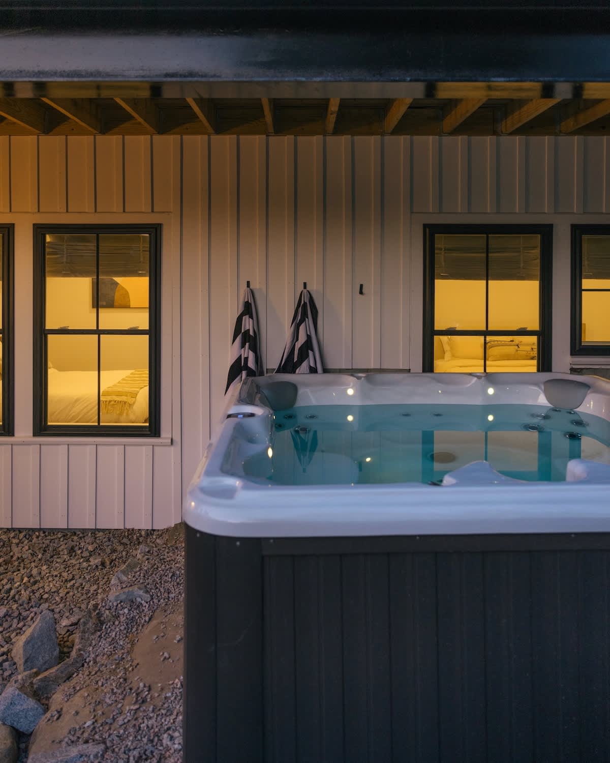 Outdoor hot tub with illuminated water, set on a gravel patio next to a white board-and-batten house with multiple windows showing warm interior lights. Two striped towels hang on the wall.
