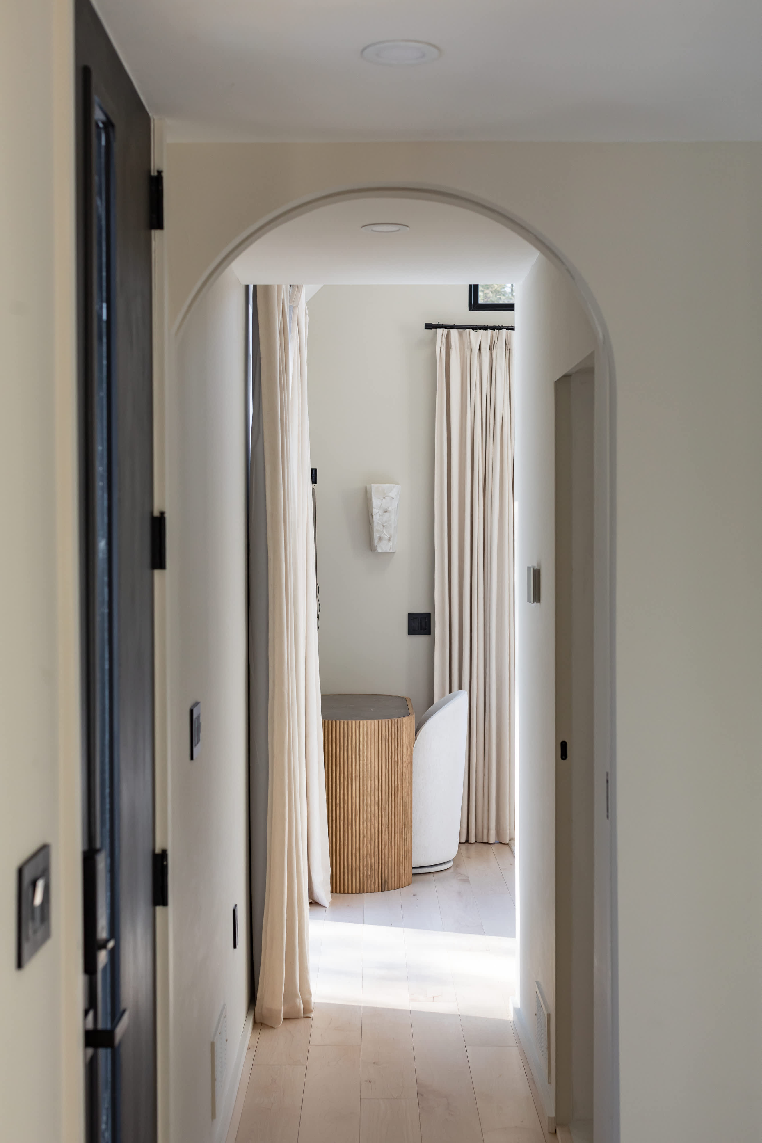 A bright hallway with light wood floors and white walls, featuring an arched doorway leading to a room with a fluted wooden side table, a white armchair, and light-colored curtains.