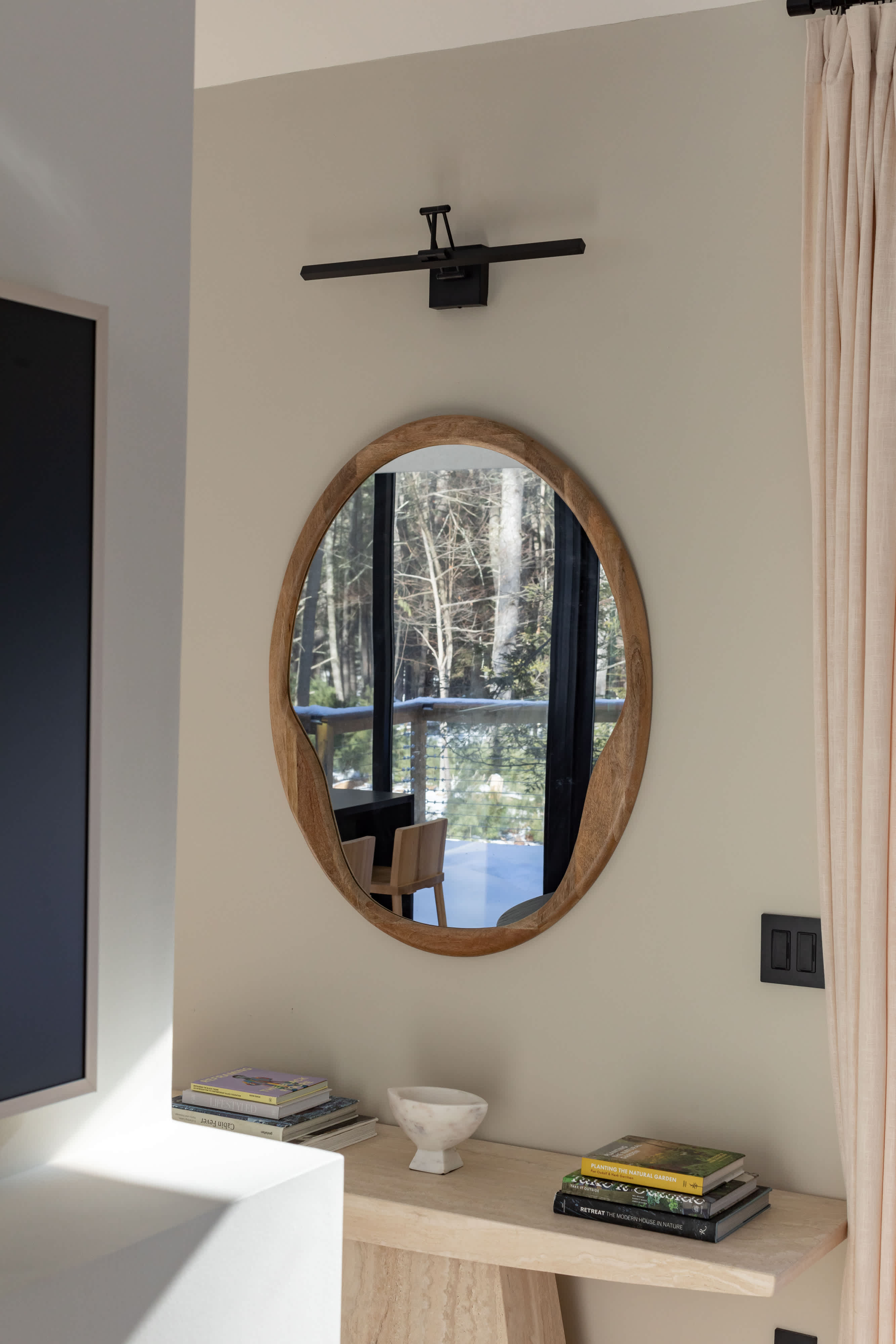 A light-colored wall features a round, organic-shaped wooden mirror hanging above a minimalist console table. Books and a decorative bowl are neatly arranged on the table, with a black picture light mounted above the mirror.