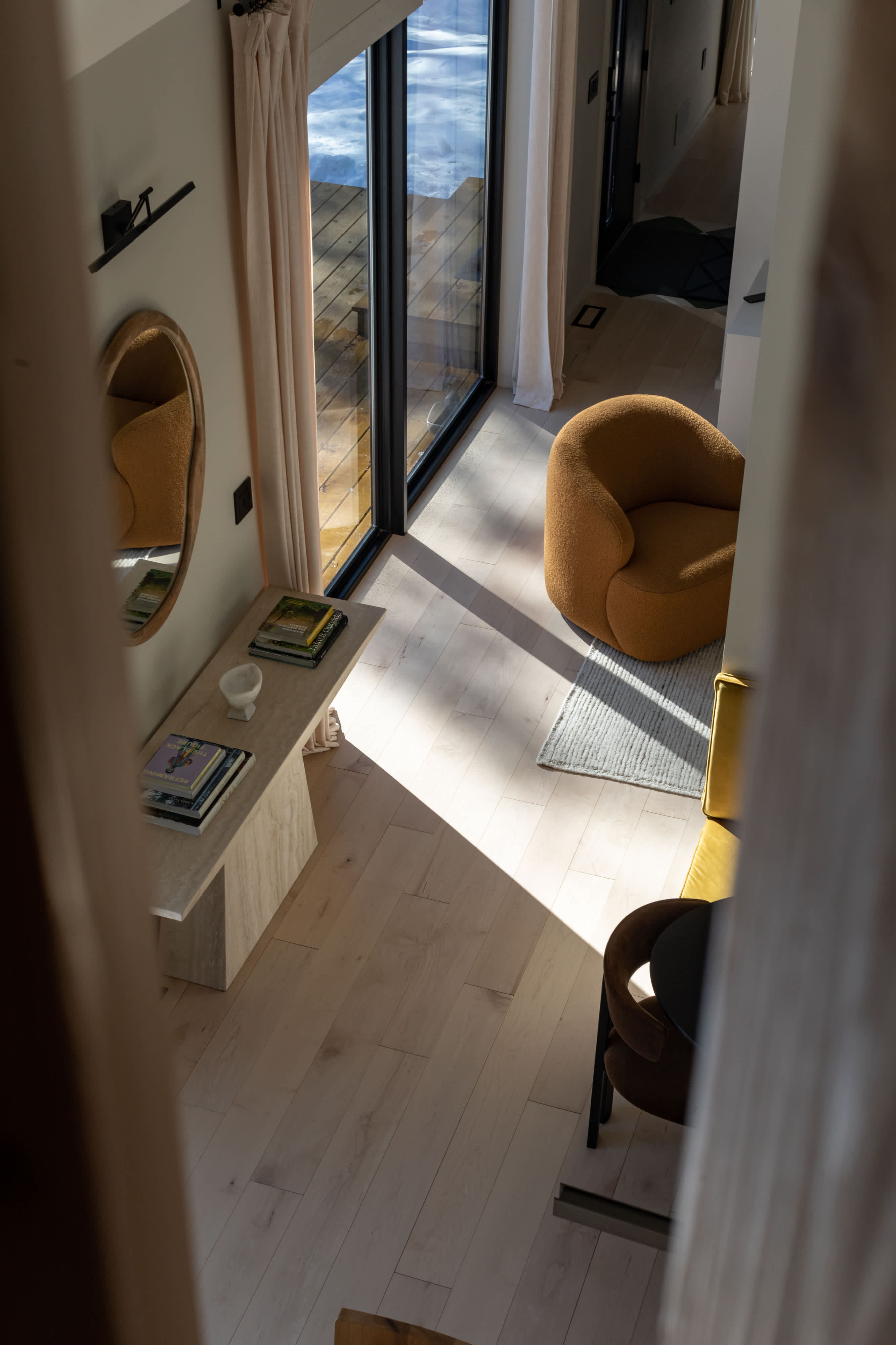 Overhead view of a bright living area with light wood floors, featuring an orange boucle armchair, a console table with books, and large glass doors that let in abundant natural light.