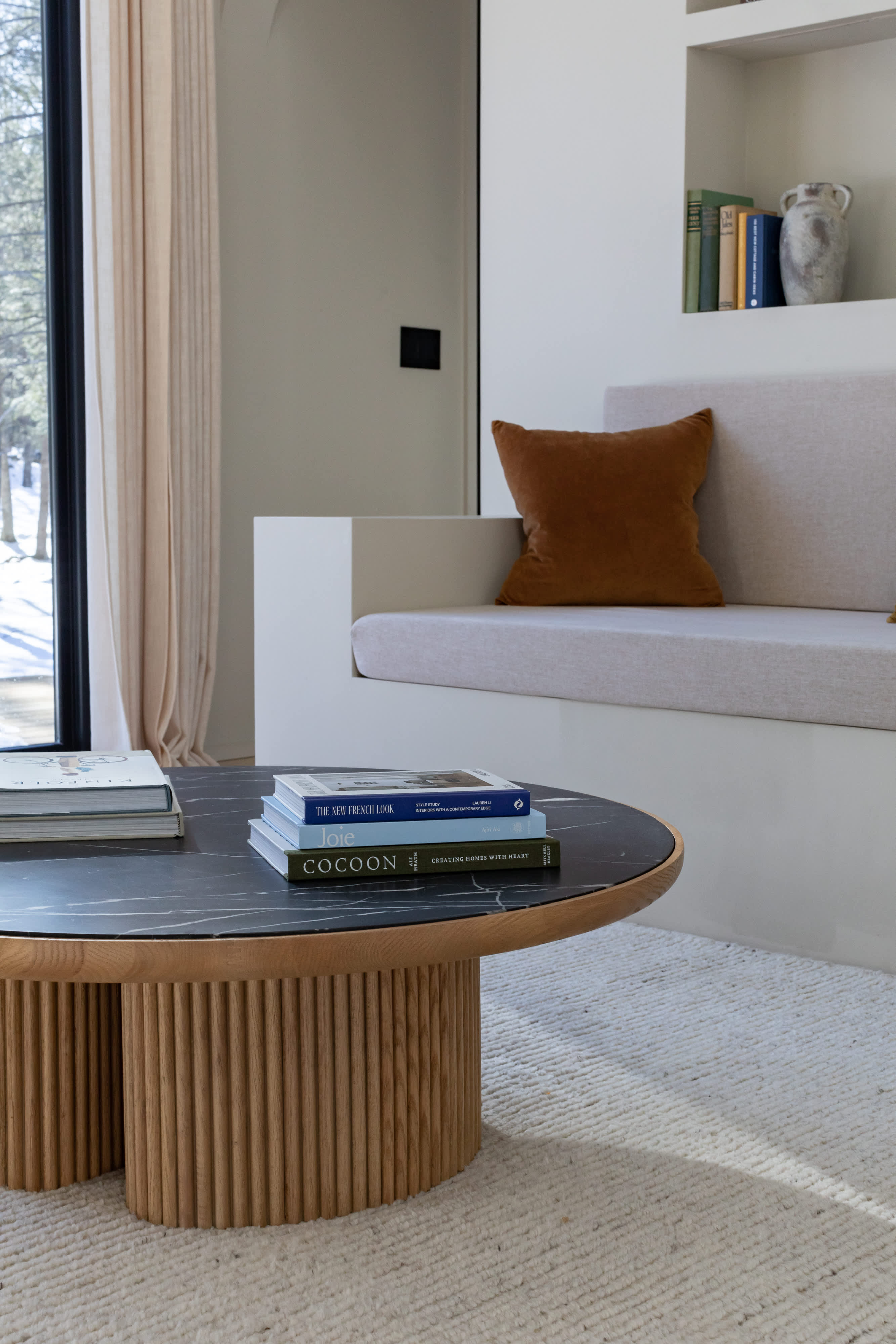 A close-up of a modern living room featuring a round coffee table with a black marble top and a fluted wooden base, adorned with books. A built-in sofa with a rust-colored cushion and a textured rug are also visible.