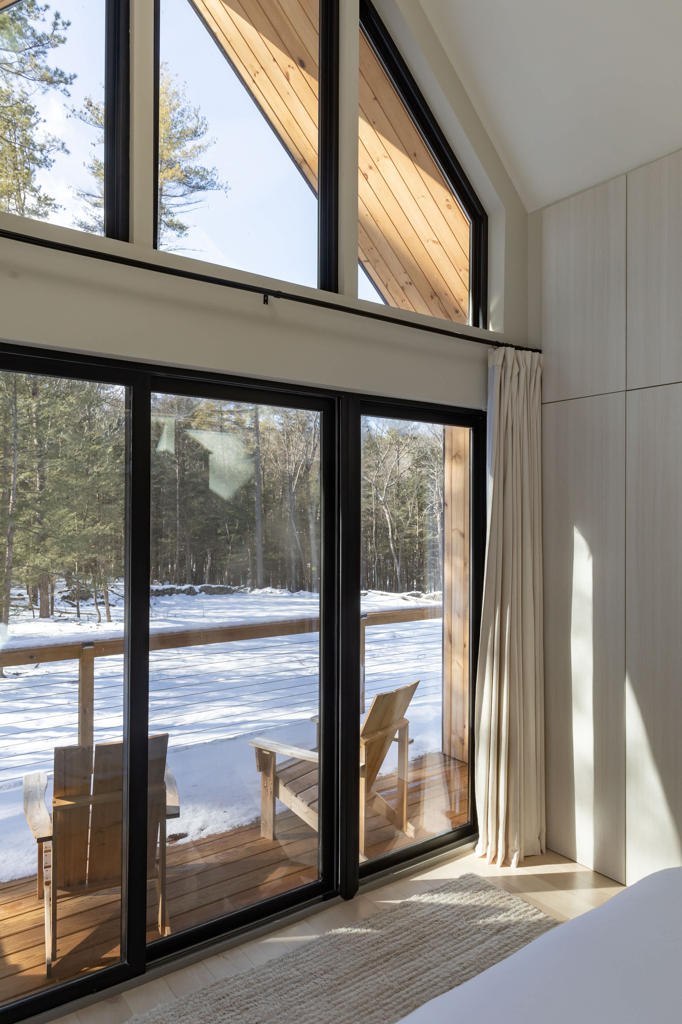 A spacious wooden deck with two Adirondack chairs, overlooking a snow-covered forest, accessed through large black-framed sliding glass doors from an interior with light wood walls and a textured rug.