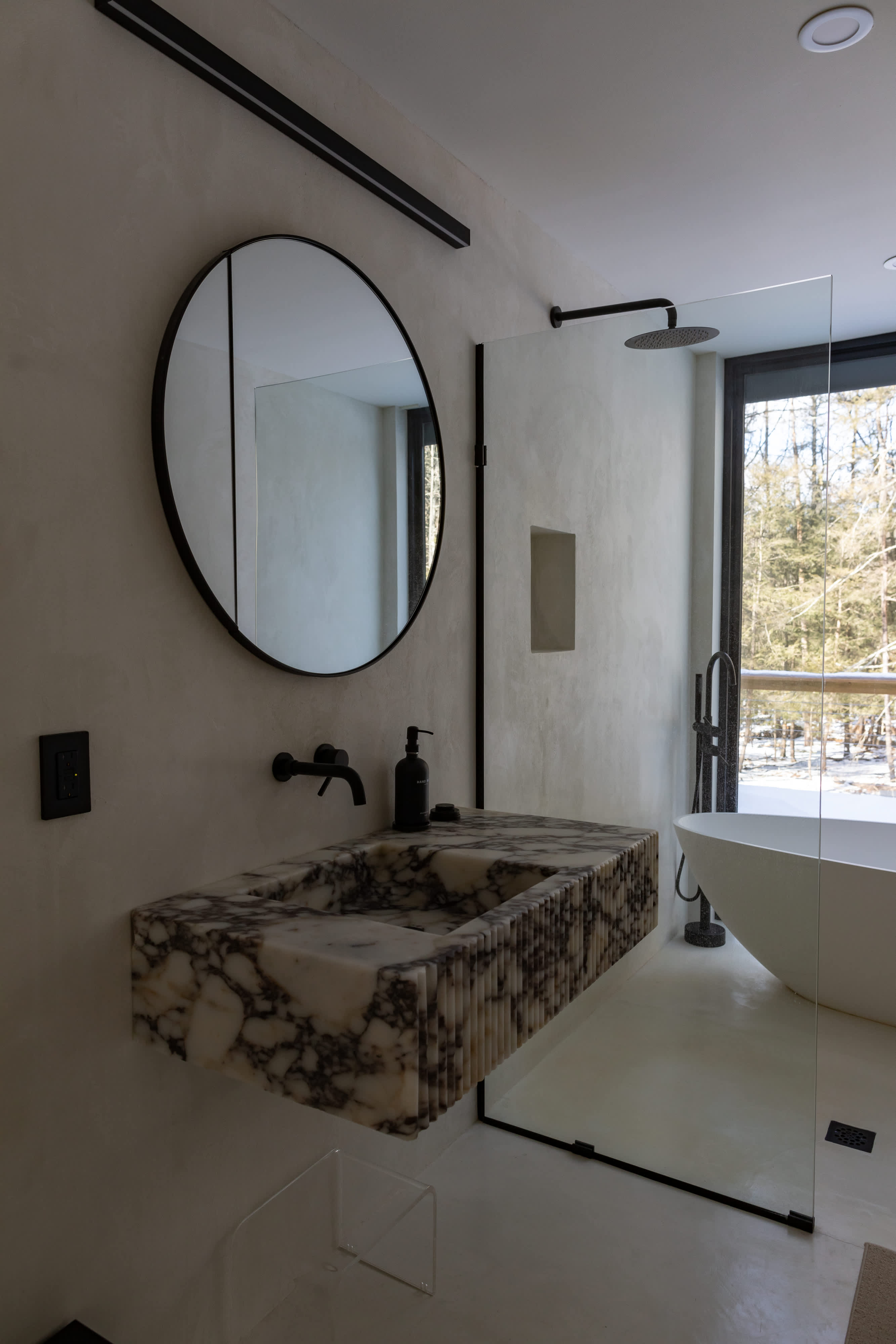 A modern bathroom featuring a unique fluted marble floating vanity with a black faucet, a large round mirror, and a glass-enclosed shower next to a freestanding tub, all with views of a snowy forest.