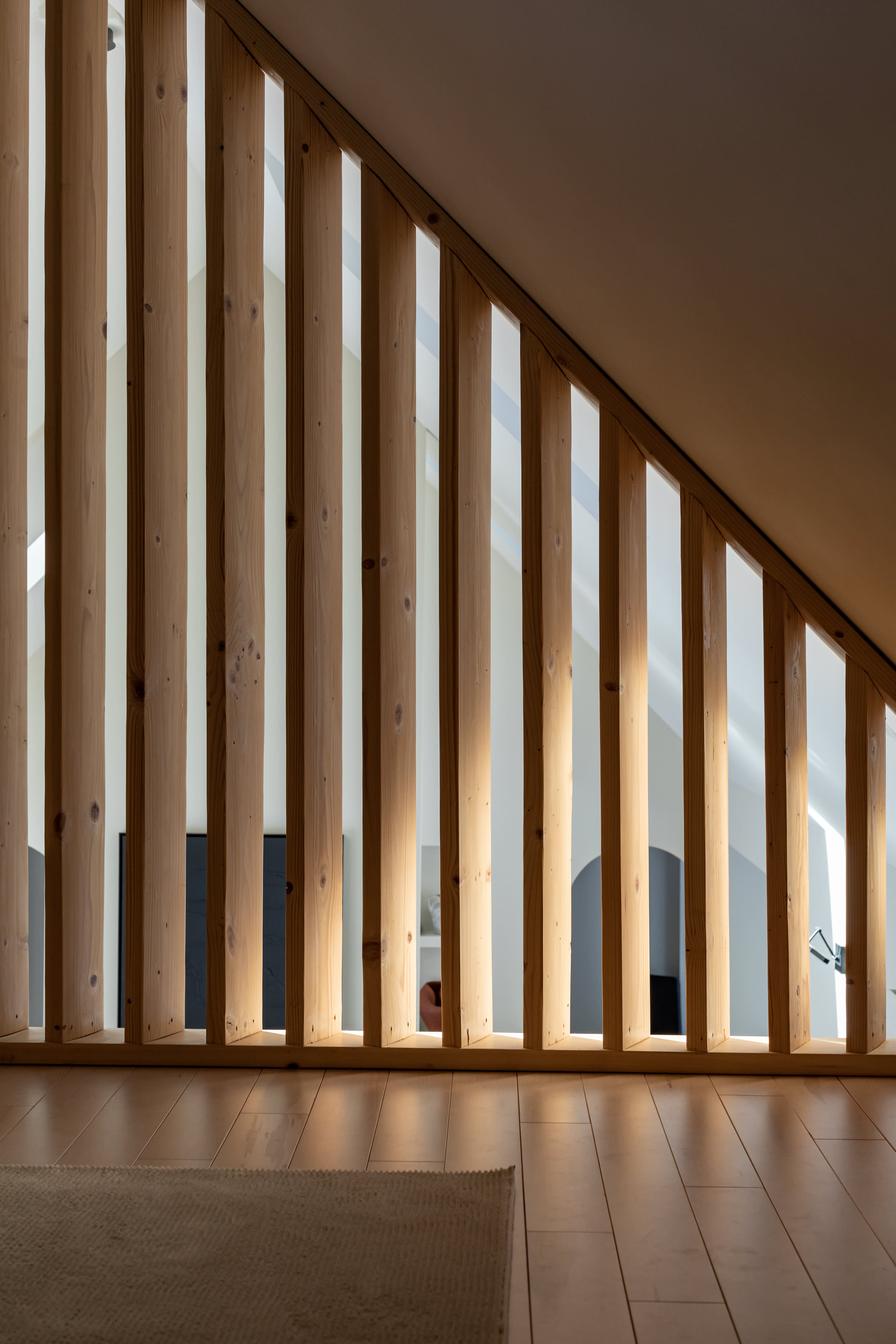 A close-up view highlights a striking wooden slat partition, creating a dynamic interplay of light and shadow on the light wood flooring. The slatted design offers partial views into an adjacent attic space.