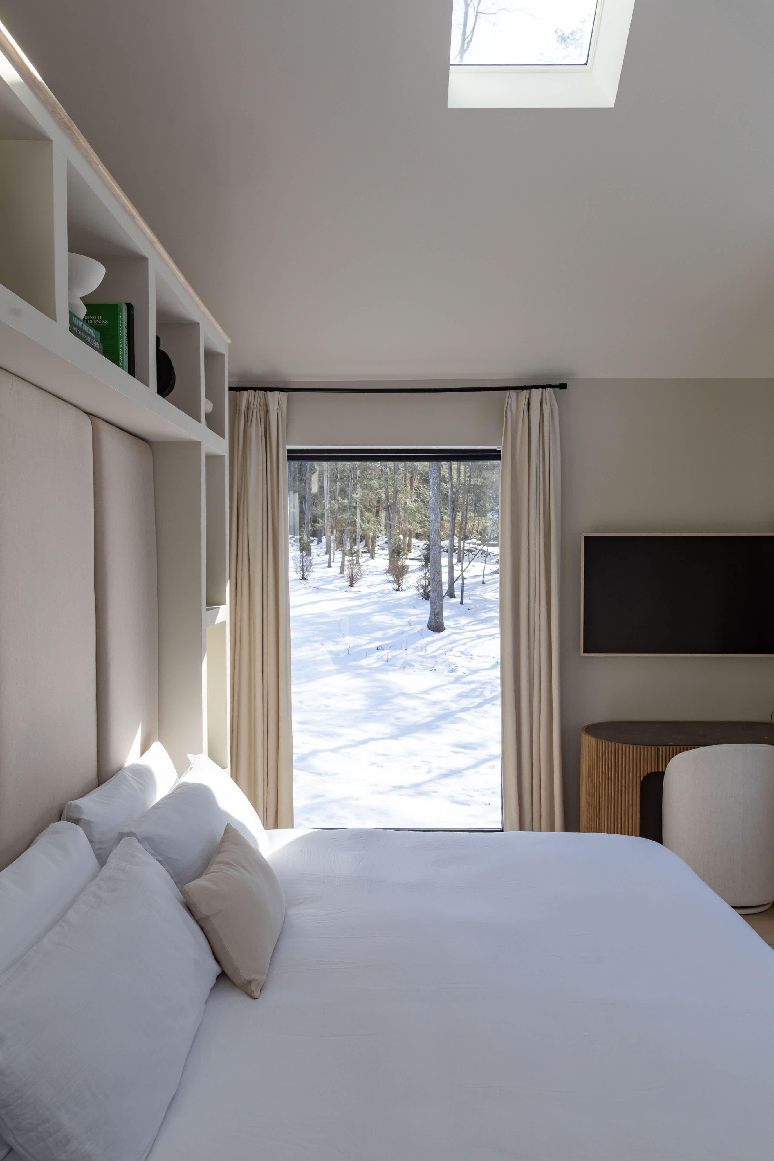 A bright bedroom with a white-linened bed facing a large window that offers a serene view of a snow-covered forest, complemented by light wood floors and a fluted wooden side table.