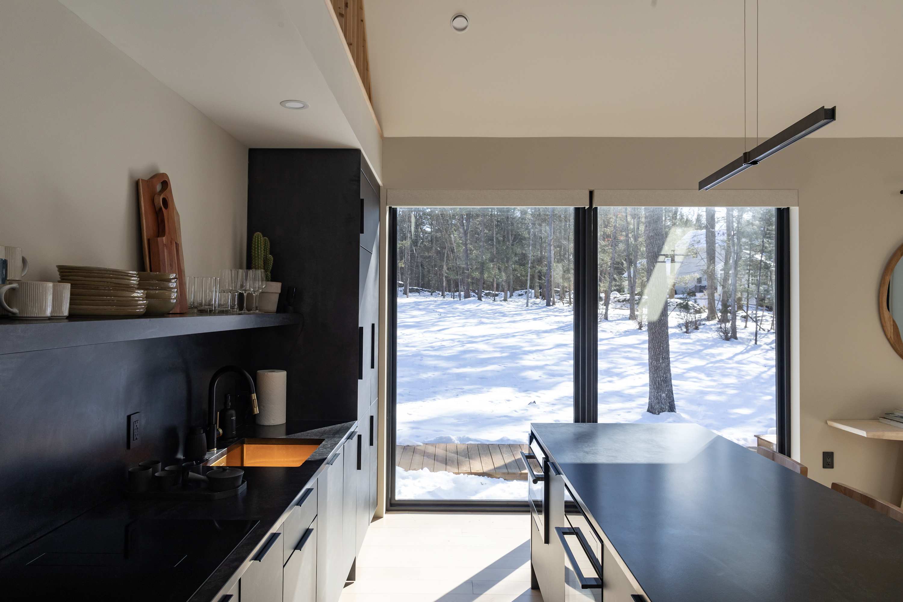 A sleek, modern kitchen features dark cabinetry, black countertops, and a large island with an integrated cooktop. A floor-to-ceiling window offers expansive views of a snowy, wooded landscape, bringing natural light and scenery into the space.