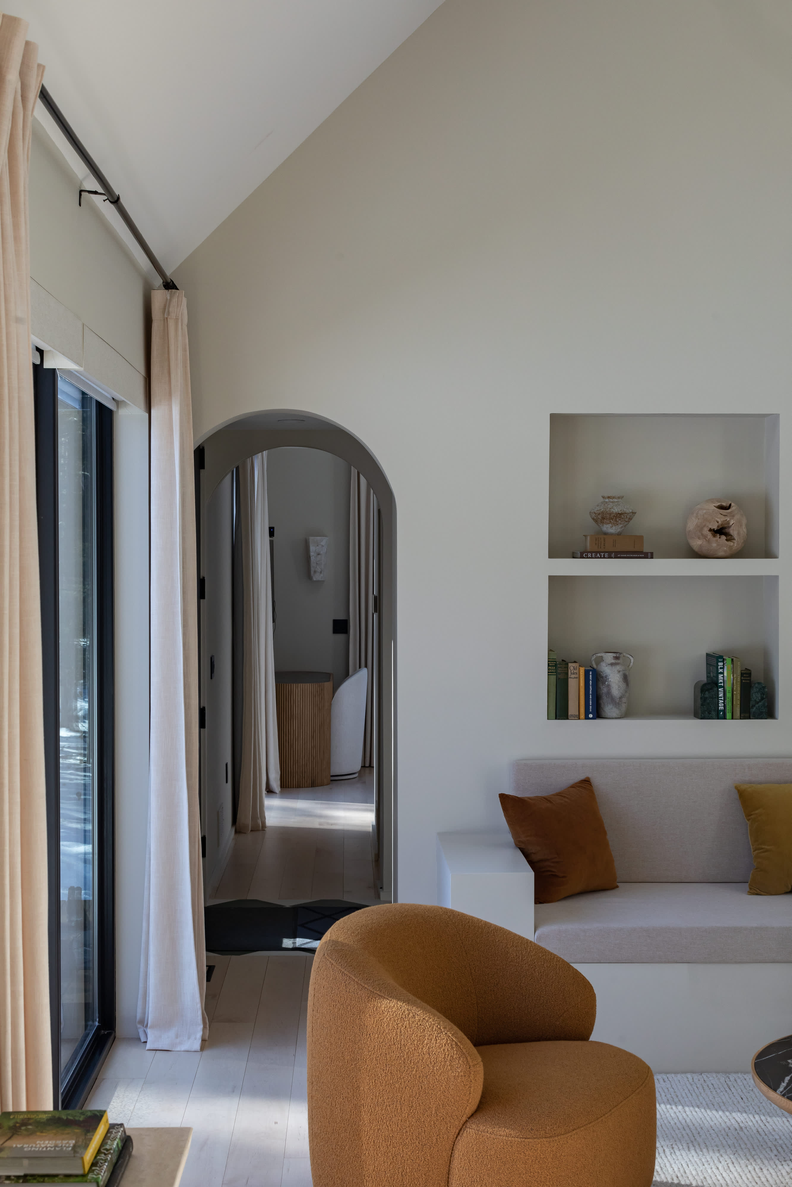 Inviting living space with a built-in light beige sofa and an orange boucle armchair, featuring light wood floors and an arched doorway leading to another part of the home. Large glass doors open to the outside.