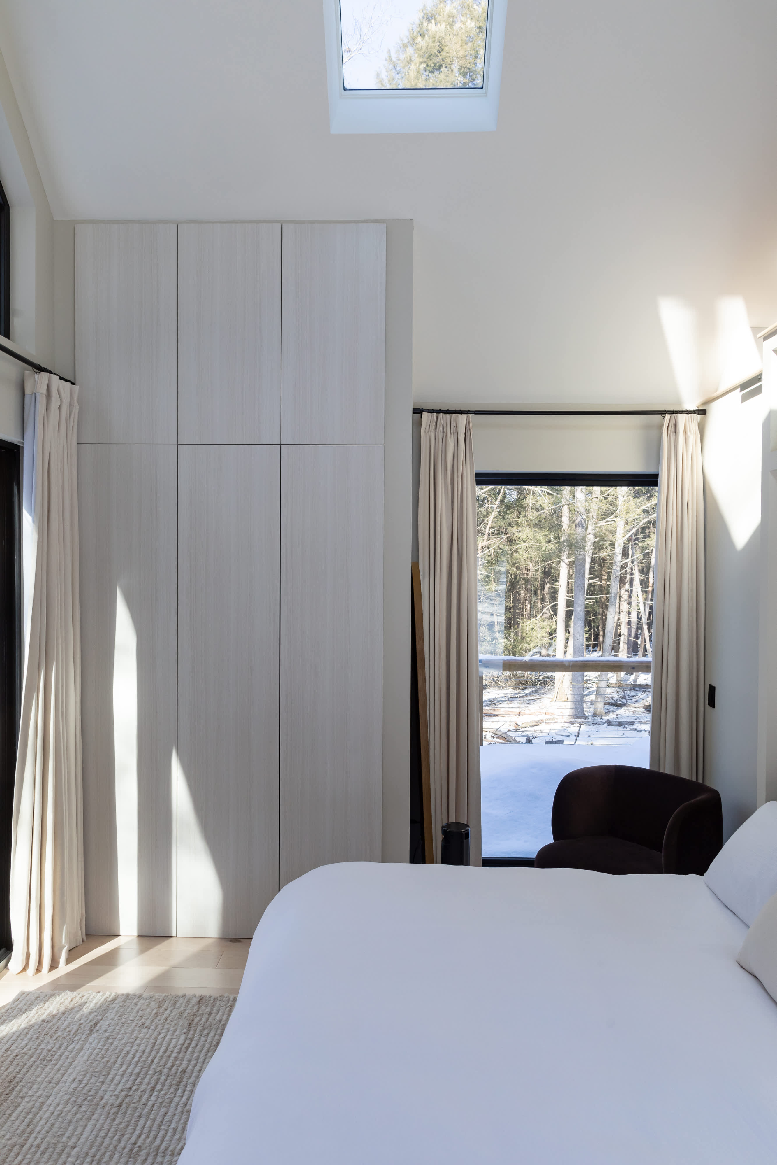 A bright bedroom featuring a white-linened bed, a large light wood wardrobe, and a dark armchair, with natural light streaming through windows that reveal a snowy outdoor landscape.