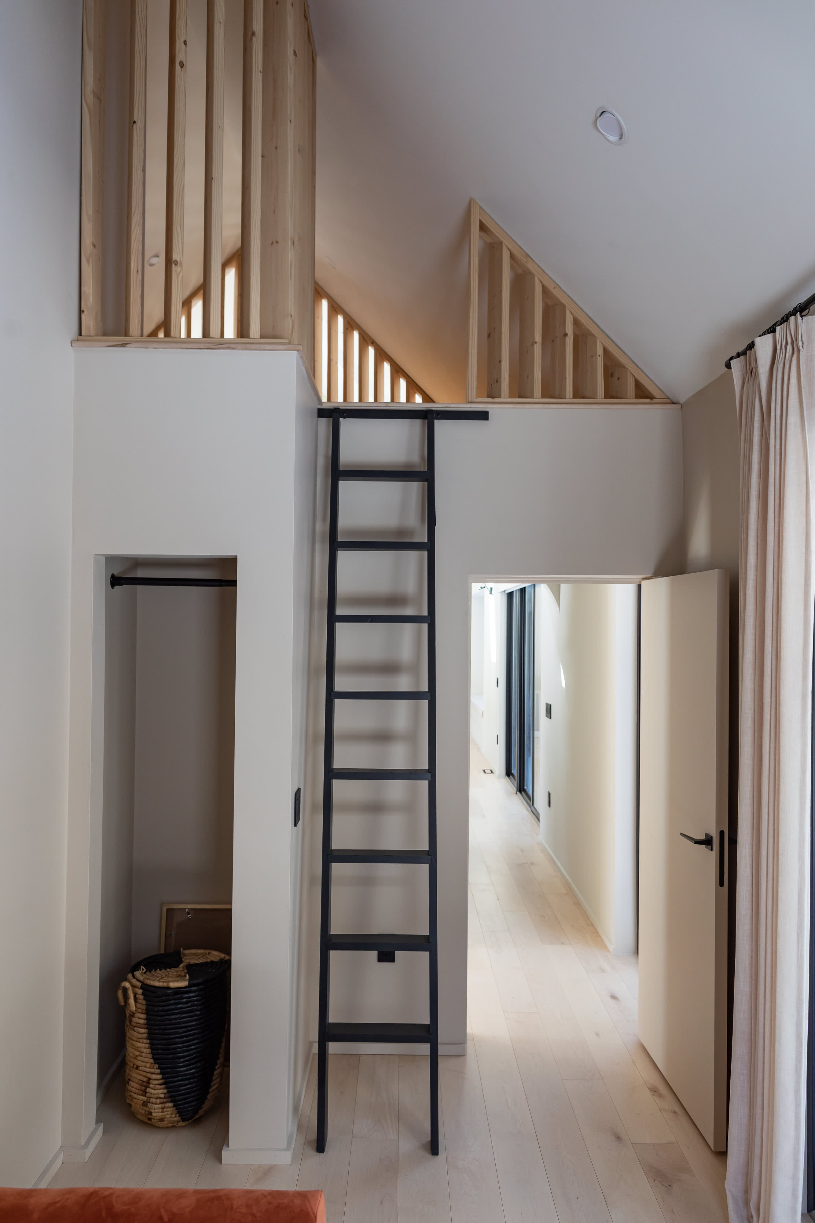 A hallway with light wood floors and white walls, featuring a black ladder leading to a wooden-railed loft space, with a glimpse into an adjacent room with an orange daybed and gallery wall.