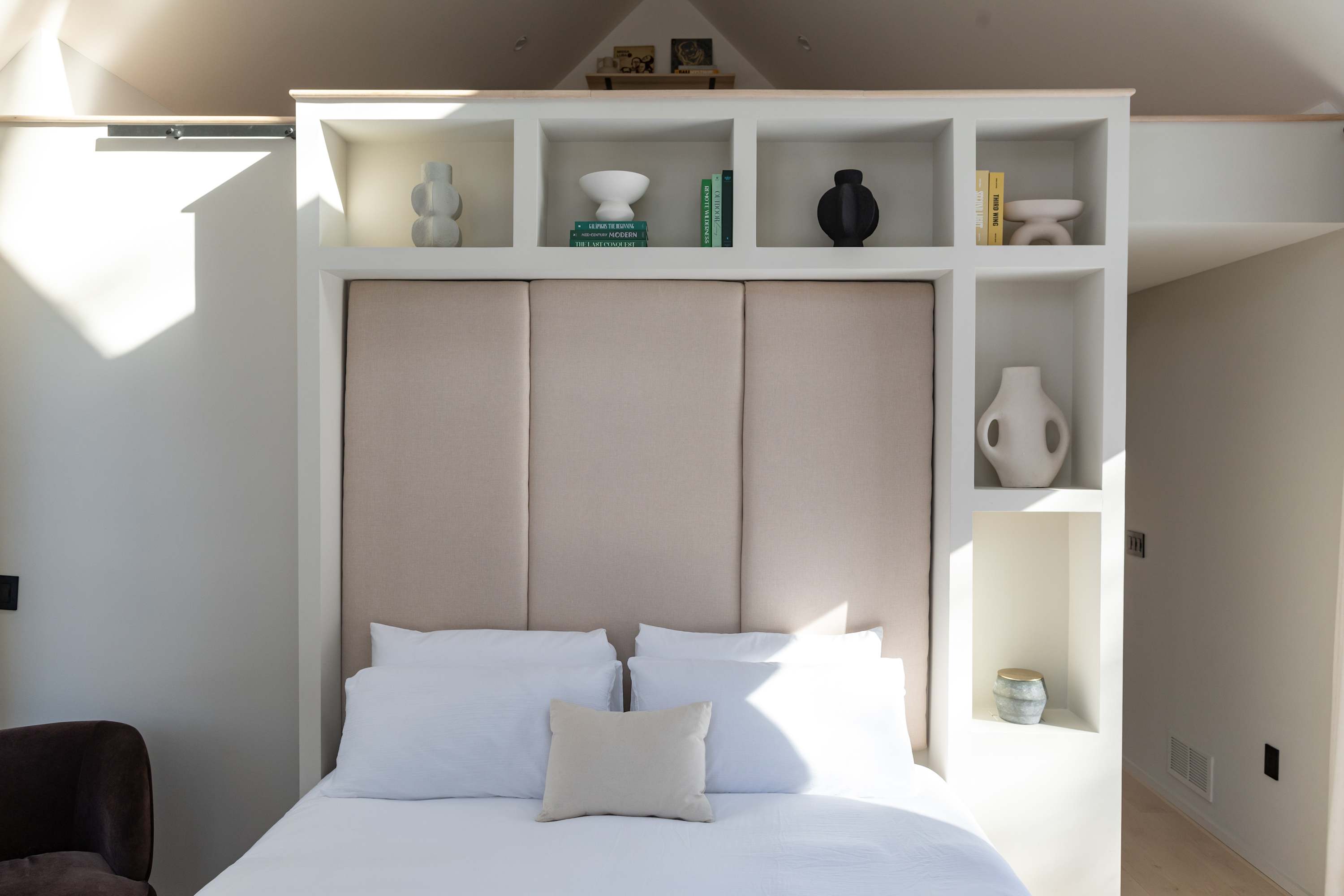 A cozy bedroom featuring a bed with crisp white linens and pillows, a beige upholstered headboard, and built-in white shelving adorned with decorative vases and books, bathed in natural light.