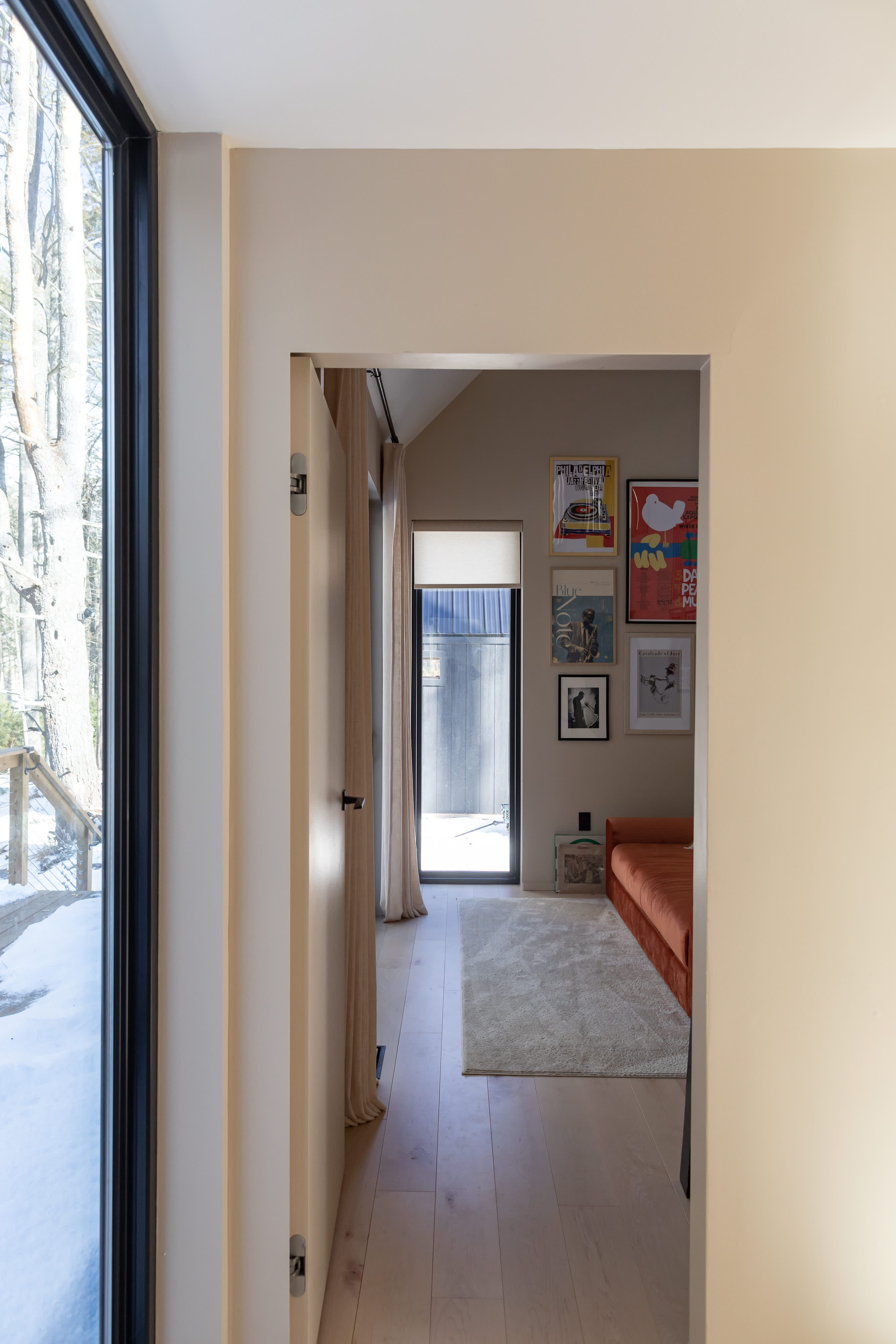 A bright hallway with light wood floors, leading into a room adorned with a gallery wall of colorful framed posters and a long orange upholstered bench, with a large window offering an outdoor view.
