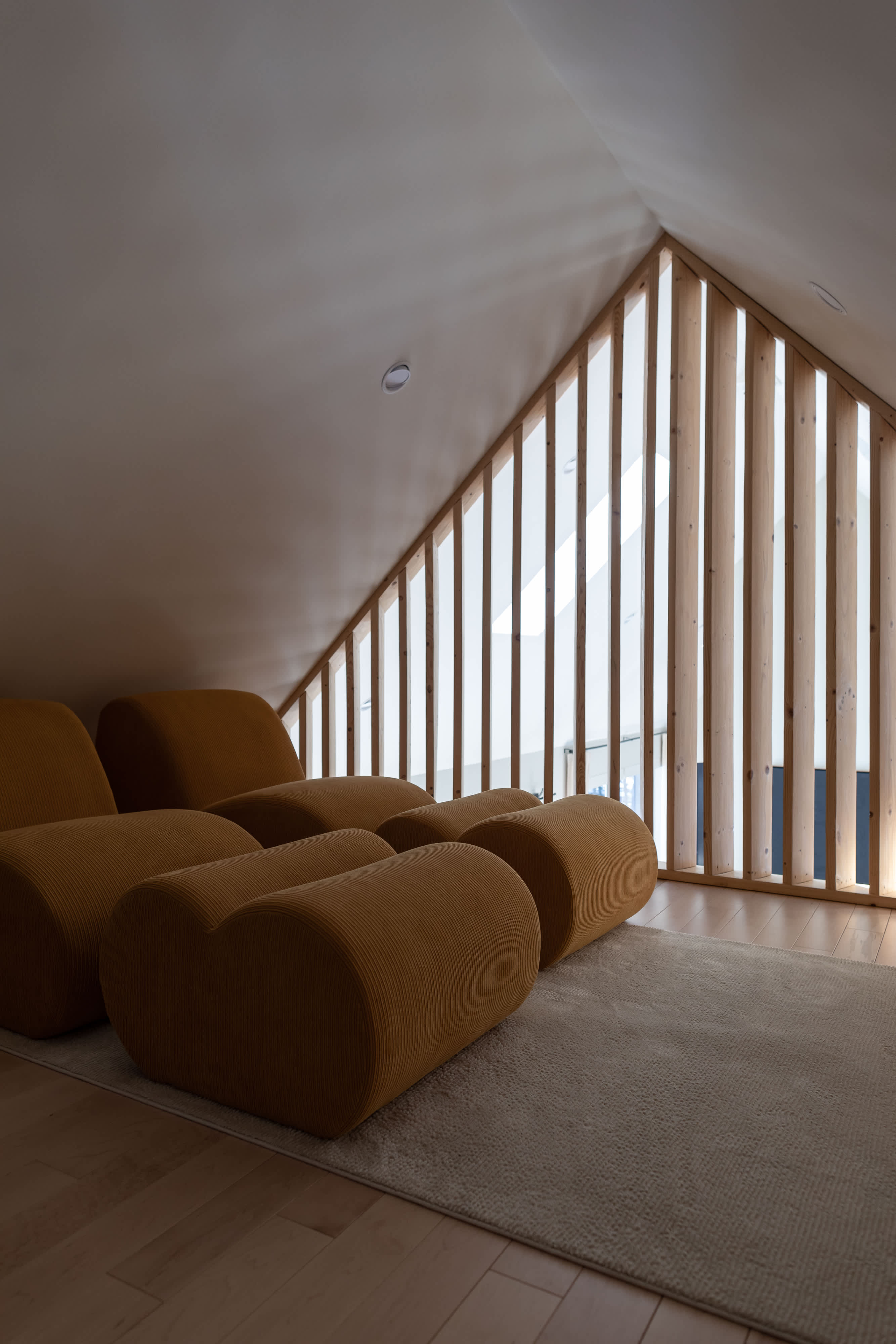 An inviting attic lounge features sloped ceilings, light wood flooring, and a unique wooden slat partition. Modular, mustard-colored upholstered seating pieces are arranged on a light rug, creating a cozy relaxation space.