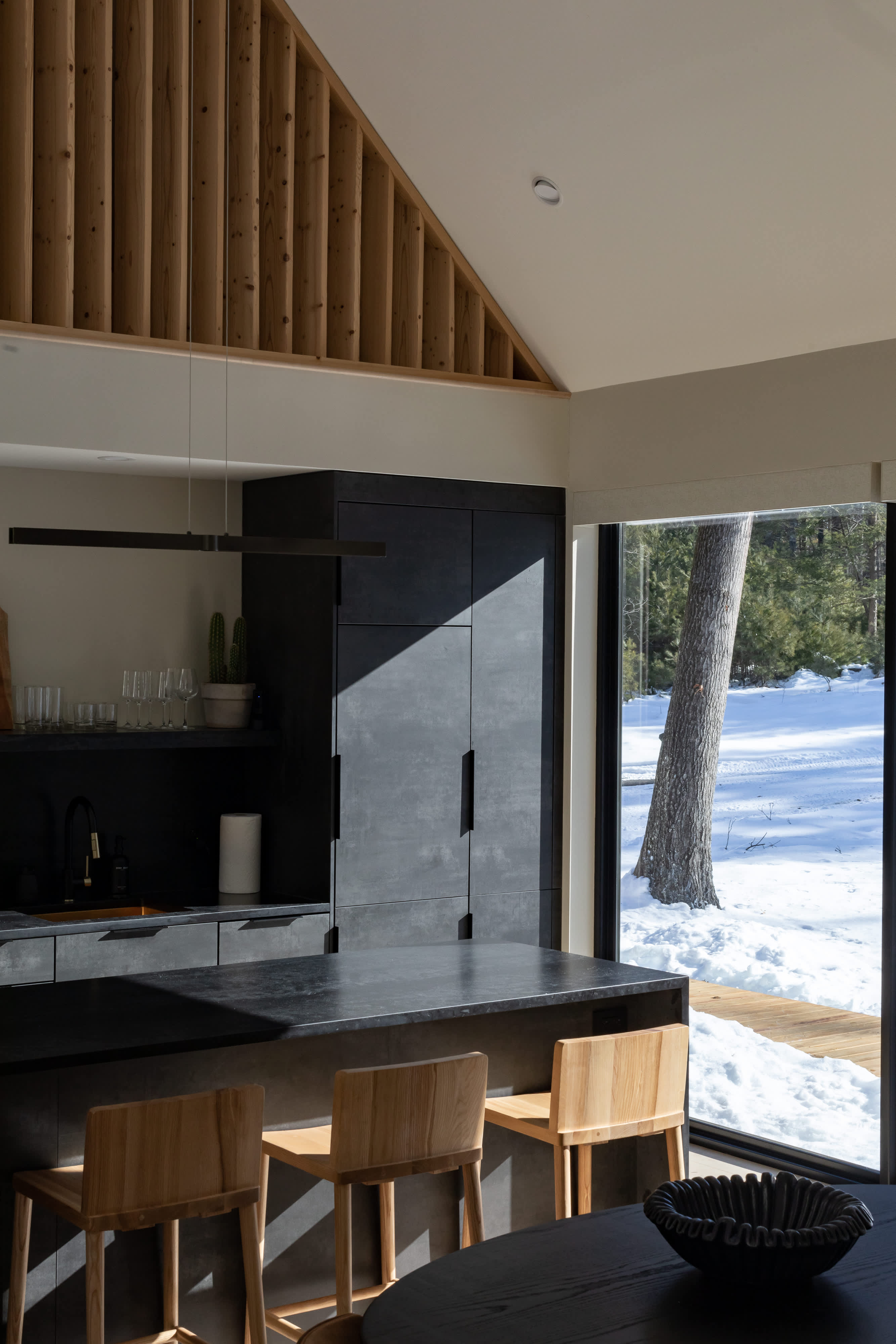 A contemporary kitchen with dark cabinetry and a large island, featuring wooden bar stools and a view through expansive windows to a snow-covered outdoor landscape. Sunlight streams in, creating strong contrasts on the dark surfaces.